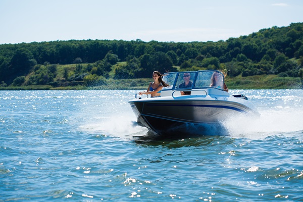 Three people riding in a speedboat on a sunny day, creating a splash as they move quickly across a lake with a forested shoreline in the background.