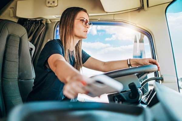 Female truck driver reaching for a smartphone while steering a commercial vehicle, illustrating distracted driving behind the wheel of a large truck.
