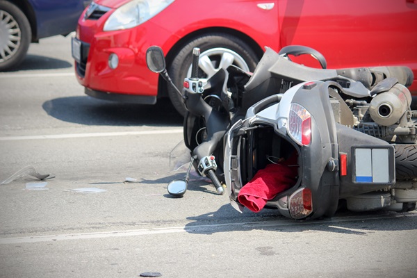 Motorcycle lying on its side in the middle of the road after a crash, with shattered debris scattered nearby and a red car parked close behind it.