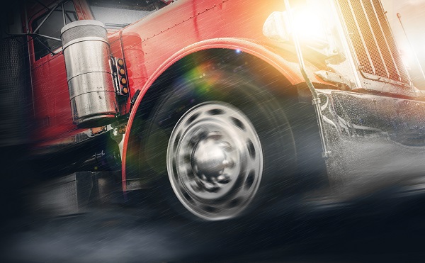 Close-up of a red semi-truck speeding through heavy rain with its front wheel in motion and headlights shining brightly.