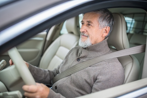 An older man with a grey beard wearing a brown cardigan and a seatbelt while driving a car.